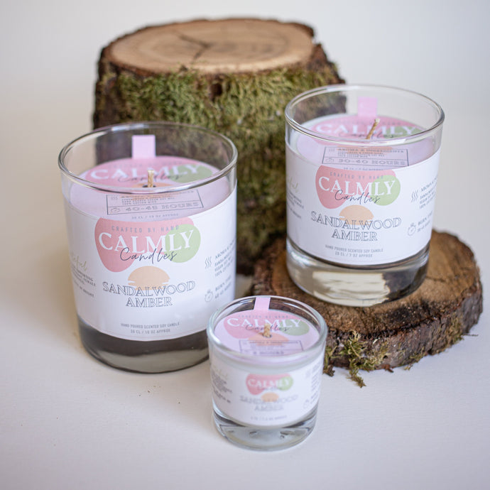 30cl, 20cl & 4cl Sandalwood & Amber Scented Soy Candle. Shown together as group stood on chopped wooden logs, with green moss on wood bark. Calmly Candles Surrey based female owned independent local candle company. 